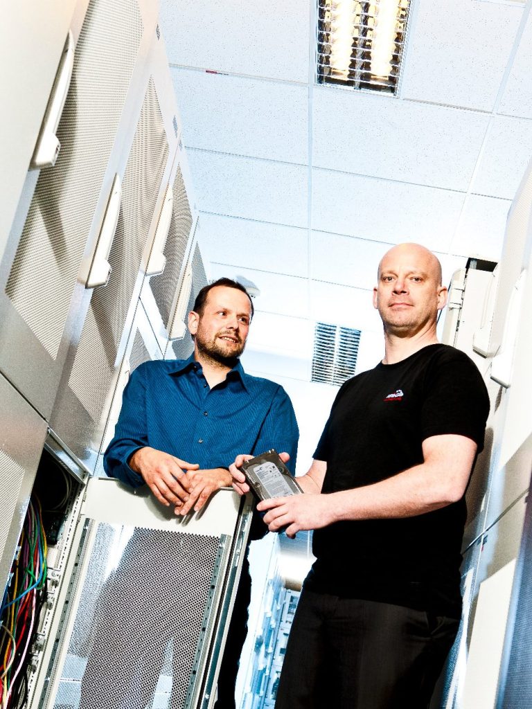 Attingo Datenrettungs-Techniker im Serverraum Zwei Männer stehen in einem Serverraum, einer hält eine Festplatte.